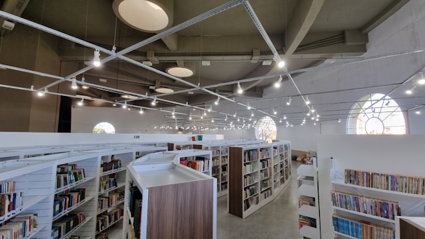 A spacious library with modern architecture features multiple rows of bookshelves filled with various books. The ceiling is high with unique circular designs and numerous pendant lights hanging above. Large arched windows allow natural light to enter, creating a bright and inviting atmosphere.