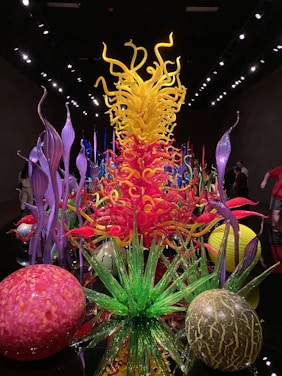 A vibrant display of blown glass sculptures featuring swirling shapes and a mix of colors. The centerpiece is a towering structure with yellow and orange tendrils surrounded by various organic forms, including green spiky plants and spherical objects in red and pink hues. The art pieces are set against a dark backdrop, creating a striking contrast.