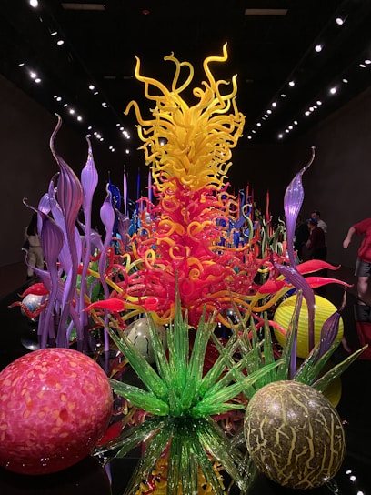 A vibrant display of blown glass sculptures featuring swirling shapes and a mix of colors. The centerpiece is a towering structure with yellow and orange tendrils surrounded by various organic forms, including green spiky plants and spherical objects in red and pink hues. The art pieces are set against a dark backdrop, creating a striking contrast.