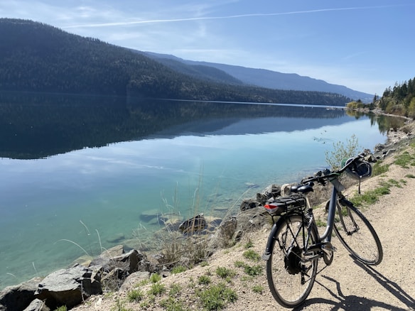 A tranquil lakeside scene features a bicycle parked on a gravel path. Clear, calm water reflects the surrounding forested mountains under a bright blue sky. Sparse vegetation and rocks edge the path, adding natural texture to the scene.