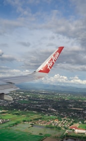 An airplane wing with a red tip featuring the AirAsia logo is prominently visible against a backdrop of partly cloudy skies. Below, a landscape of lush green fields, scattered buildings, and a few patches of water stretches into the distance.