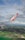 An airplane wing with a red tip featuring the AirAsia logo is prominently visible against a backdrop of partly cloudy skies. Below, a landscape of lush green fields, scattered buildings, and a few patches of water stretches into the distance.