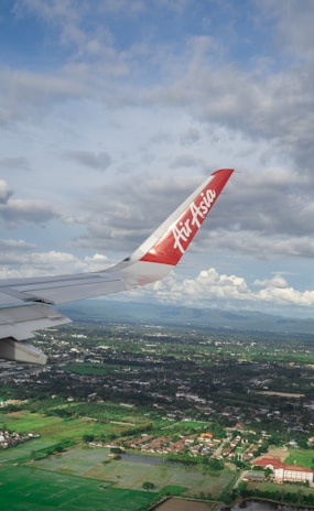 An airplane wing with a red tip featuring the AirAsia logo is prominently visible against a backdrop of partly cloudy skies. Below, a landscape of lush green fields, scattered buildings, and a few patches of water stretches into the distance.