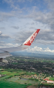 An airplane wing with a red tip featuring the AirAsia logo is prominently visible against a backdrop of partly cloudy skies. Below, a landscape of lush green fields, scattered buildings, and a few patches of water stretches into the distance.