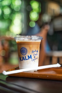 A plastic cup filled with a layered iced coffee beverage, marked with the words 'KALM Art Craft Culture' against a blurred, bokeh-style background. The drink rests on a wooden tray with a white straw placed beside it.
