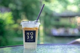 A plastic cup filled with an iced coffee drink, topped with a layer of cream. The cup bears the text NO 39 CAFE & YAKINIKU and sits on a glass table. The background is a blurred natural green setting, creating a tranquil outdoor atmosphere.