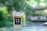A plastic cup filled with an iced coffee drink, topped with a layer of cream. The cup bears the text NO 39 CAFE & YAKINIKU and sits on a glass table. The background is a blurred natural green setting, creating a tranquil outdoor atmosphere.