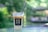 A plastic cup filled with an iced coffee drink, topped with a layer of cream. The cup bears the text NO 39 CAFE & YAKINIKU and sits on a glass table. The background is a blurred natural green setting, creating a tranquil outdoor atmosphere.