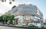 A modern shopping mall with a unique, lattice-designed facade and large glass windows. The building is marked with the name 'MAYA' in large letters on the exterior. It features a busy street in front with cars, motorbikes, and pedestrians. There are trees and a small structure resembling traditional architecture near the entrance.