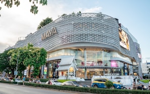 A modern shopping mall with a unique, lattice-designed facade and large glass windows. The building is marked with the name 'MAYA' in large letters on the exterior. It features a busy street in front with cars, motorbikes, and pedestrians. There are trees and a small structure resembling traditional architecture near the entrance.