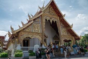 An intricately designed temple with ornate golden decorations on its roof and facade, complemented by white pillars. The structure features steep, gilded eaves with elaborate carvings. Several people are gathered in front of the temple, including tourists taking photos and exploring the area. Lush greenery surrounds the temple, and the sky is partly cloudy, adding to the peaceful atmosphere.