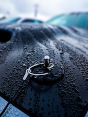 Raindrops cover the surface of a dark, metallic car hood, creating a glossy, reflective effect. In the center, a metallic fastener, reminiscent of a car hood pin, stands out with its shiny, textured surface. The background is blurred, emphasizing the focus on the fastener and the raindrops.