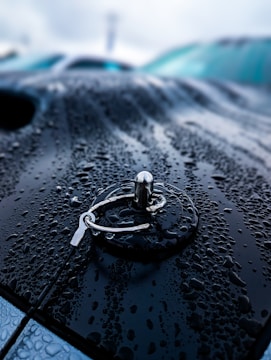 Raindrops cover the surface of a dark, metallic car hood, creating a glossy, reflective effect. In the center, a metallic fastener, reminiscent of a car hood pin, stands out with its shiny, textured surface. The background is blurred, emphasizing the focus on the fastener and the raindrops.