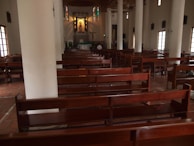 A spacious church interior featuring rows of wooden pews and large white columns. At the far end, a well-lit altar with an artwork or statue hangs below a crucifix. Stained glass windows add colorful accents to the otherwise serene and muted space.