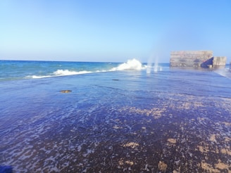 A concrete pier extends into the ocean, with waves crashing against its sides. The ocean is a deep blue, contrasting with the bright sky overhead. On the right, there are large concrete blocks that appear man-made, possibly part of a structure or breakwater.