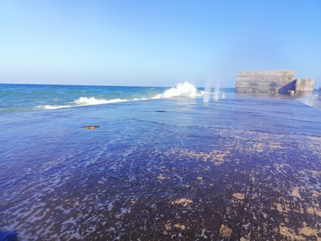 A concrete pier extends into the ocean, with waves crashing against its sides. The ocean is a deep blue, contrasting with the bright sky overhead. On the right, there are large concrete blocks that appear man-made, possibly part of a structure or breakwater.