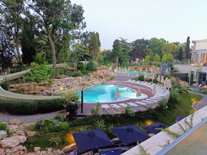 A beautiful outdoor swimming pool surrounded by lush greenery and stone landscaping. Numerous sun loungers with closed umbrellas line the poolside. A mix of trees and shrubs provide a serene backdrop, enhancing the natural beauty. The scene is well-lit, likely suggesting a luxury resort or spa setting.