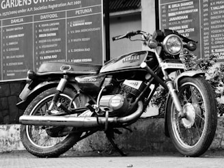 A classic Yamaha RX100 motorcycle parked against a wall with a large signboard in the background. The motorcycle appears to be in good condition, showcasing its iconic design and branding. It is parked on a tiled pavement, leaning slightly on its kickstand. The signboard lists names and appears to be associated with an organization or residential complex.