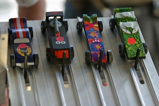 Four colorful, small model cars are positioned on a metallic race track. Each car has a distinct design: one with red and white stripes, another with a black body and flame graphics, a third with a vibrant, multi-colored floral pattern, and the last one with green scales and cartoonish eyes. The track is designed with lanes, and metal rods are holding the cars in place.