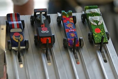 Four colorful, small model cars are positioned on a metallic race track. Each car has a distinct design: one with red and white stripes, another with a black body and flame graphics, a third with a vibrant, multi-colored floral pattern, and the last one with green scales and cartoonish eyes. The track is designed with lanes, and metal rods are holding the cars in place.