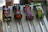 Four colorful, small model cars are positioned on a metallic race track. Each car has a distinct design: one with red and white stripes, another with a black body and flame graphics, a third with a vibrant, multi-colored floral pattern, and the last one with green scales and cartoonish eyes. The track is designed with lanes, and metal rods are holding the cars in place.