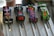 Four colorful, small model cars are positioned on a metallic race track. Each car has a distinct design: one with red and white stripes, another with a black body and flame graphics, a third with a vibrant, multi-colored floral pattern, and the last one with green scales and cartoonish eyes. The track is designed with lanes, and metal rods are holding the cars in place.
