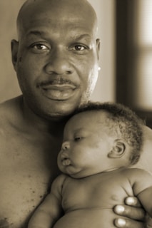 A man holding a baby close, exuding warmth and tenderness. The baby rests peacefully in his arms, showcasing a serene moment. Both are shirtless, emphasizing a theme of natural bonding.