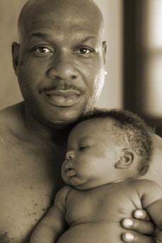 A man holding a baby close, exuding warmth and tenderness. The baby rests peacefully in his arms, showcasing a serene moment. Both are shirtless, emphasizing a theme of natural bonding.