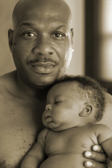 A man holding a baby close, exuding warmth and tenderness. The baby rests peacefully in his arms, showcasing a serene moment. Both are shirtless, emphasizing a theme of natural bonding.