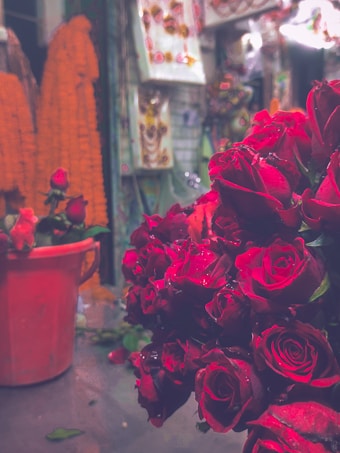 A vibrant bunch of red roses is clustered in the foreground, glistening with droplets of water. To the left, a bucket holds more roses and an orange garland hangs prominently. The background features a variety of decorative items with bright colors and intricate designs, likely indicating a shop or market setting.