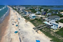 Aerial view of a long stretch of beach lined with beach umbrellas and vacationers. On one side, there are waves crashing onto the shore and on the other, a row of beachfront properties featuring large houses with decks and swimming pools. Beyond the houses, there are marshlands and more residential buildings in the distance.