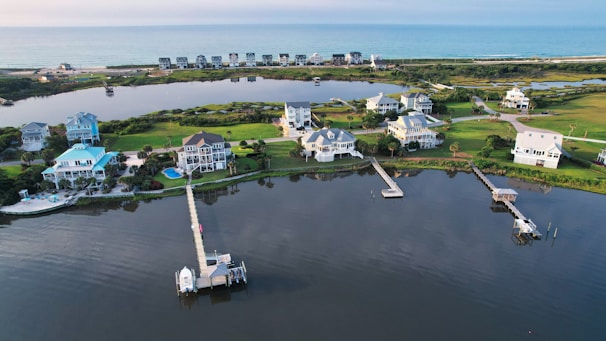 An aerial view of a coastal residential area featuring several large, well-maintained houses with docks extending into a calm body of water. The landscape includes neatly trimmed lawns and a mix of light and dark green vegetation. The area is bordered by a stretch of ocean in the background, giving a serene and picturesque setting.