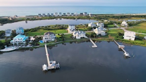 An aerial view of a coastal residential area featuring several large, well-maintained houses with docks extending into a calm body of water. The landscape includes neatly trimmed lawns and a mix of light and dark green vegetation. The area is bordered by a stretch of ocean in the background, giving a serene and picturesque setting.