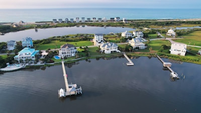 An aerial view of a coastal residential area featuring several large, well-maintained houses with docks extending into a calm body of water. The landscape includes neatly trimmed lawns and a mix of light and dark green vegetation. The area is bordered by a stretch of ocean in the background, giving a serene and picturesque setting.