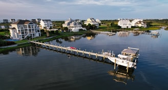 Several upscale waterfront homes are situated along a tranquil waterway, with a long pier extending into the water. There are boats docked at the pier, and the scene reflects a peaceful coastal setting.