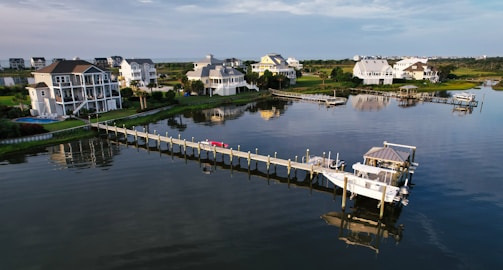 Several upscale waterfront homes are situated along a tranquil waterway, with a long pier extending into the water. There are boats docked at the pier, and the scene reflects a peaceful coastal setting.