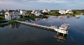 Several upscale waterfront homes are situated along a tranquil waterway, with a long pier extending into the water. There are boats docked at the pier, and the scene reflects a peaceful coastal setting.