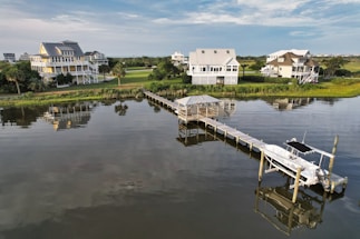A scenic view of several waterfront homes surrounded by lush greenery. The calm water reflects the houses, pier, and sky. A long wooden pier extends into the water, featuring a small gazebo and a docked boat.