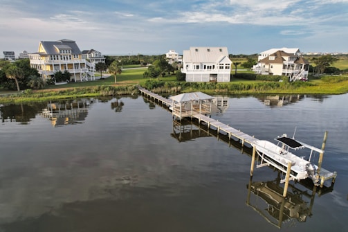 A scenic view of several waterfront homes surrounded by lush greenery. The calm water reflects the houses, pier, and sky. A long wooden pier extends into the water, featuring a small gazebo and a docked boat.