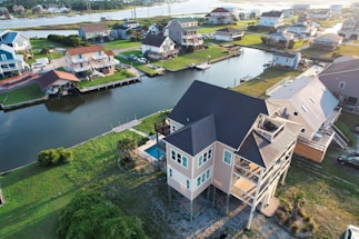Aerial view of a suburban neighborhood with several houses situated along a waterway. The homes appear elevated, possibly due to being in a flood-prone area. There are grassy lawns and docks extending into the canal. The area is well-maintained and showcases a mix of modern and traditional architecture.