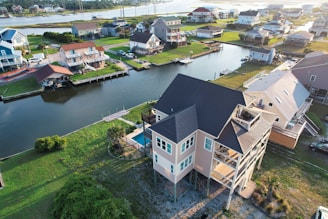 Aerial view of a suburban neighborhood with several houses situated along a waterway. The homes appear elevated, possibly due to being in a flood-prone area. There are grassy lawns and docks extending into the canal. The area is well-maintained and showcases a mix of modern and traditional architecture.