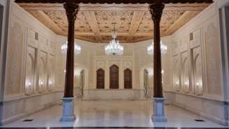 An ornate interior features intricate wooden ceilings with detailed carvings and patterns. The walls are adorned with decorative designs and columns, and elegant chandeliers hang from the ceiling. Symmetrical arches and a blend of warm colors create a luxurious atmosphere.