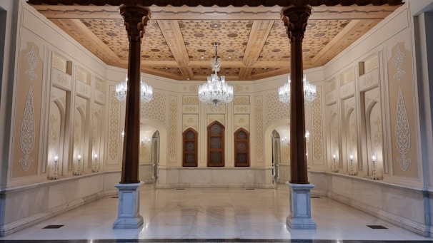 An ornate interior features intricate wooden ceilings with detailed carvings and patterns. The walls are adorned with decorative designs and columns, and elegant chandeliers hang from the ceiling. Symmetrical arches and a blend of warm colors create a luxurious atmosphere.