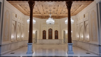 An ornate interior features intricate wooden ceilings with detailed carvings and patterns. The walls are adorned with decorative designs and columns, and elegant chandeliers hang from the ceiling. Symmetrical arches and a blend of warm colors create a luxurious atmosphere.
