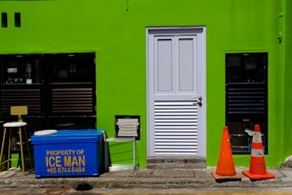 A bright green wall features a white door in the center. To the left of the door are two black-framed windows, a stool, and a blue cooler with the text 'Property of ICE MAN' and a phone number written in yellow. On the right side of the door, there are two orange traffic cones placed on the ground. There is also a rectangular vent near the bottom left corner of the door.