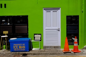 A bright green wall features a white door in the center. To the left of the door are two black-framed windows, a stool, and a blue cooler with the text 'Property of ICE MAN' and a phone number written in yellow. On the right side of the door, there are two orange traffic cones placed on the ground. There is also a rectangular vent near the bottom left corner of the door.