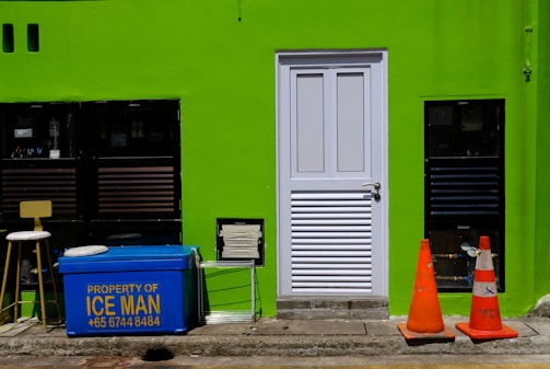 A bright green wall features a white door in the center. To the left of the door are two black-framed windows, a stool, and a blue cooler with the text 'Property of ICE MAN' and a phone number written in yellow. On the right side of the door, there are two orange traffic cones placed on the ground. There is also a rectangular vent near the bottom left corner of the door.