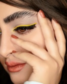 A close-up of a person's face showcasing intricate makeup. The eye makeup features bold, yellow eyeliner along the upper lash line and thick, black lashes. Eyebrows are groomed and dark. Lips are painted a matte terracotta color, matching the fingernail polish on the hand that gently touches the face.