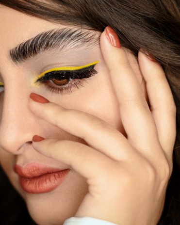 A close-up of a person's face showcasing intricate makeup. The eye makeup features bold, yellow eyeliner along the upper lash line and thick, black lashes. Eyebrows are groomed and dark. Lips are painted a matte terracotta color, matching the fingernail polish on the hand that gently touches the face.