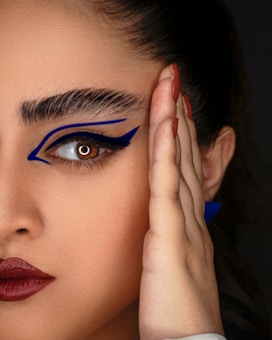A close-up image of a person's face focusing on the eye with bold, artistic blue eyeliner extending towards the temple. The eyebrows are thick and prominent, and the lips are painted with a deep red lipstick. One hand is gently placed against the side of the face, showcasing neatly manicured nails with a terracotta shade.